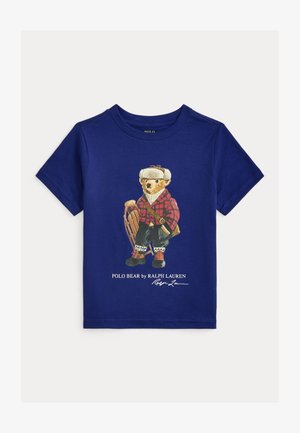 Tumši zila T-krekls ar lāča attēlu, kas valkā sarkanu rūtainu jaku, sniega zābakus un tur ragavas, kā arī ar tekstu "Polo Bear by Ralph Lauren".