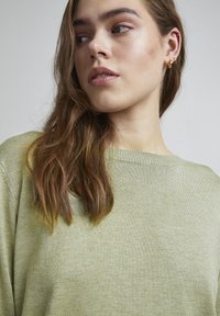 Šviesiai žalia megzta palaidinė su plačiu apykaklės iškirpimu. Minkšta tekstūra ir laisvas siluetas. Modelis turi ilgus plaukus ir vilki mažas auksines auskarų kilpas.