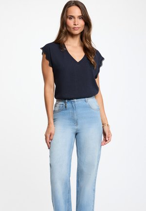 Kvinde iført navyblå V-hals ærmeløs top med bølgede kanter og lyseblå high-waist jeans, stående mod en hvid baggrund.