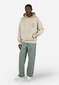 Sweatshirt bege com capuz e um bolso frontal, combinado com jeans azul claro que apresentam um design lateral em listras vermelhas e brancas. Ténis brancos completam o visual.