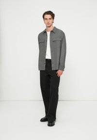 Muž stojaci opretý o bielu stenu, oblečený v sivej košeli na gombíky, bielom tričku, čiernych nohaviciach a čiernych topánkach, smerujúci dopredu s uvoľneným postojom.