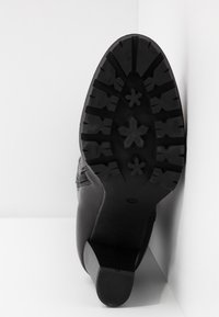 Bota negra de tobillo, parte superior de cuero, suela de goma texturizada con tacos multidireccionales y un pequeño tacón, colocada sobre un fondo blanco.