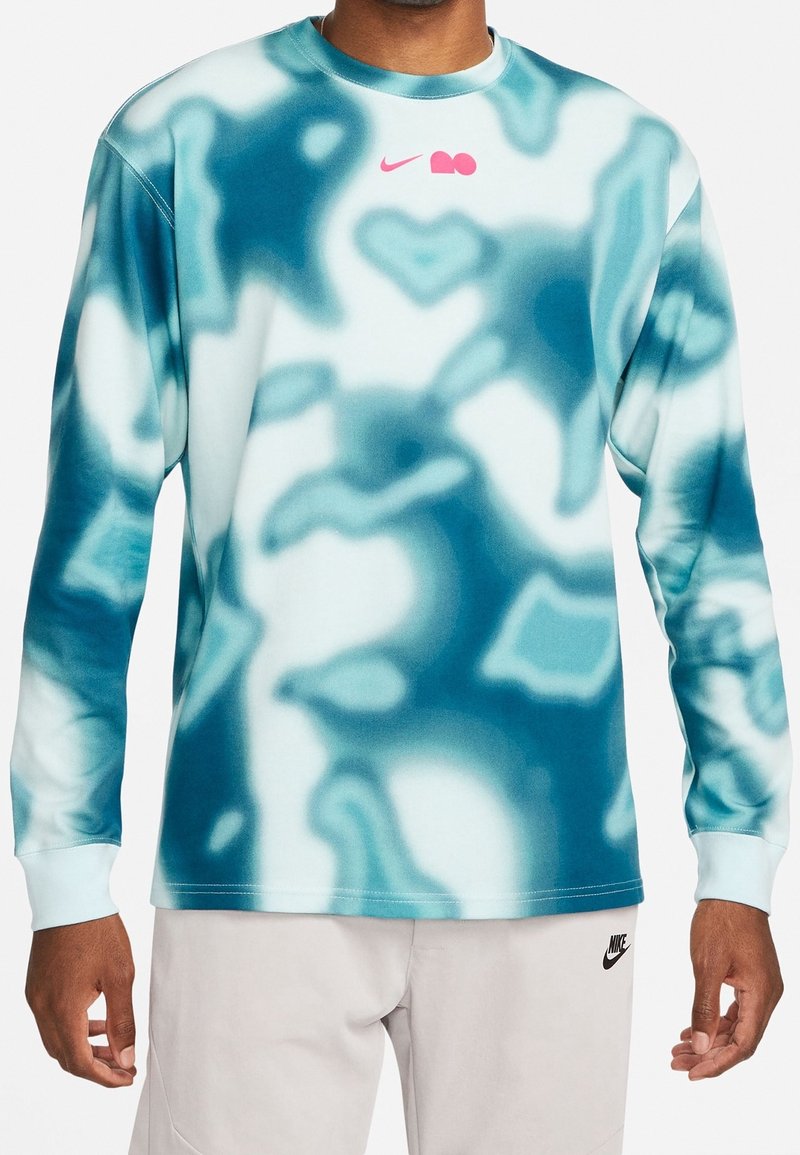 Homme portant un t-shirt à manches longues tie-dye turquoise et blanc avec un logo Nike rose, associé à un short Nike beige clair.