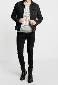 Chaqueta de mezclilla negra, camiseta gráfica gris con diseño de calavera, jeans skinny negros y botas negras con cordones. Diseño simple y ajustado en todo.