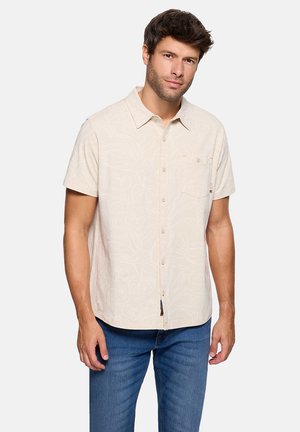 Hombre con cabello corto y oscuro, vestido con una camisa beige de manga corta con botones y patrones florales sutiles, y pantalones vaqueros azules, de pie contra un fondo liso.