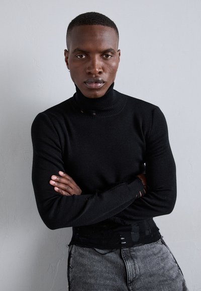 Μαύρος ζιβάγκο με ριγωτό λαιμό και ταλαιπωρημένα μανίκια, φορεμένος πάνω από γκρι ταλαιπωρημένα τζιν. Μοντέλο ποζάρει με τα χέρια σταυρωμένα.