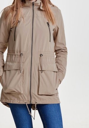 Femme portant une veste beige zippée avec quatre poches avant et des cordons, associée à un jean bleu sur un fond uni.