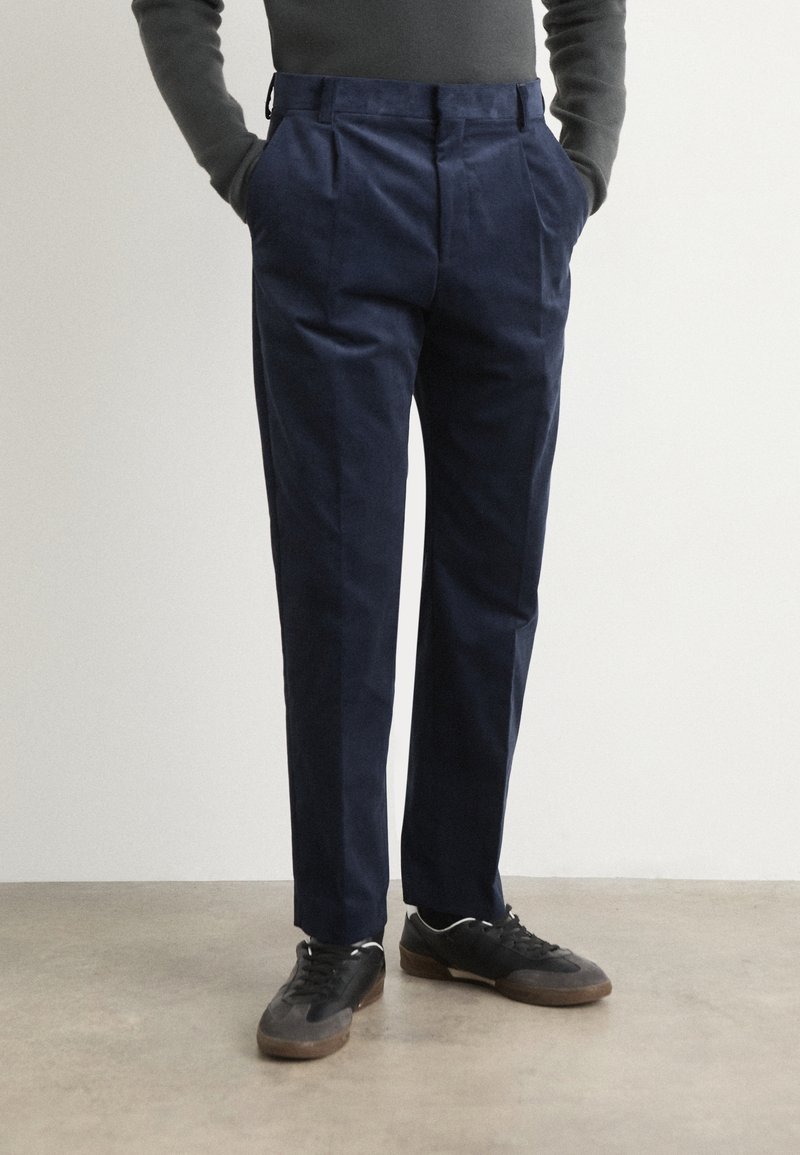 Navyblå byxor gjorda av bomullsblandat tyg; veckad design, rak ben, sidofickor och bälteshällor. I kombination med svarta sneakers.