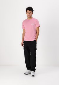 Růžové bavlněné tričko s krátkým rukávem, s bílým logem. Spárováno s černými cargo kalhotami a bílými teniskami. Jednoduchý, casual outfit.