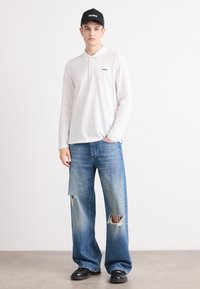 Hvit langermet polo med svart logo på venstre bryst, kombinert med lyseblå vide jeans med hull. Svart caps og sko.