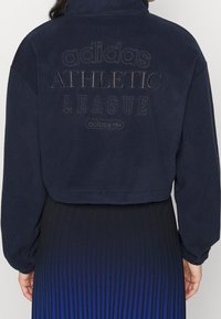 Person som bär en marinblå Adidas fleecejacka med "adidas ATHLETIC LEAGUE" broderat på ryggen och en blå plisserad kjol.