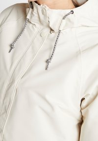 Detailní záběr osoby v světle béžové bundě s kapucí, která má přední zip a vzorované tkaničky.