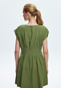 Grünes Faltenkleid mit kurzen Ärmeln, taillierter Oberteil und ausgestelltem Rock. Hergestellt aus einem strukturierten Stoff, zeigt es minimale Nähdetails.