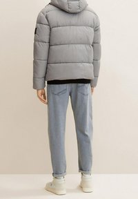 Szara pikowana kurtka z kapturem, widok od tyłu. Charakteryzuje się poziomymi przeszyciami i czarnym akcentem na lewym rękawie. W połączeniu z jasnoniebieskimi dżinsami.