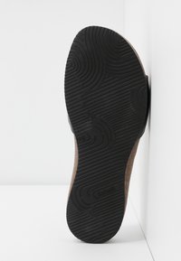 Semelle en caoutchouc noir d'une chaussure marron avec des motifs de grip texturés, positionnée verticalement contre un mur blanc en coin.