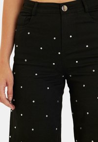Pantaloni in denim neri con vita alta, decorati con applicazioni di borchie argento sparse e chiusura con bottone frontale.