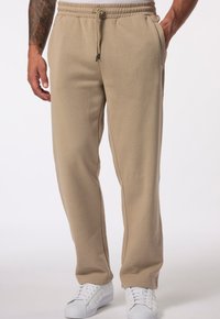 Beige tepláky vyrobené z měkkého materiálu, s elastickým pasem se šňůrkou, bočními kapsami a volným střihem. Nosí se s bílými teniskami.