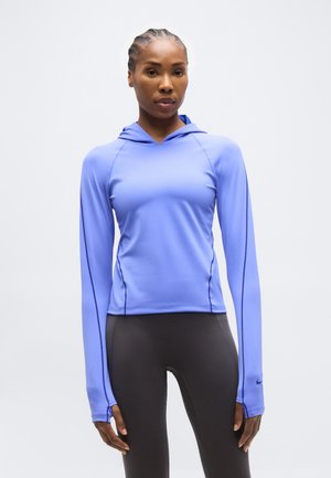 Kvinde med flettet hår iført en tætsiddende lyse lilla hættetrøje med lange ærmer og mørke leggings, stående mod en ensfarvet baggrund.