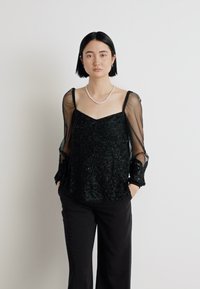 Schwarze Perlenbluse mit durchsichtigen langen Ärmeln, quadratischem Ausschnitt und aufwendigen Musterdetails, kombiniert mit schwarzen Hosen.