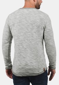 Pull gris avec tricot texturé, manches longues et encolure ronde. Présente une petite étiquette de marque en bas à gauche de l'ourlet.