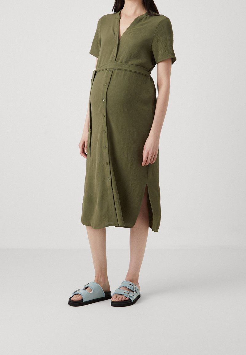 Femme enceinte en robe olive à manches courtes boutonnée sur le devant et en sandales bleu clair, debout devant un fond uni.