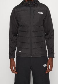 Sort dunjakke med lynlås foran, hætte og tætsiddende ærmer. Har en quiltet tekstur og The North Face-logoet på brystet.