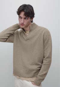 Beige strikket sweater med høj hals og ribstrikkede manchetter, der har en afslappet pasform og blød tekstur. Båret over en beige skjorte.