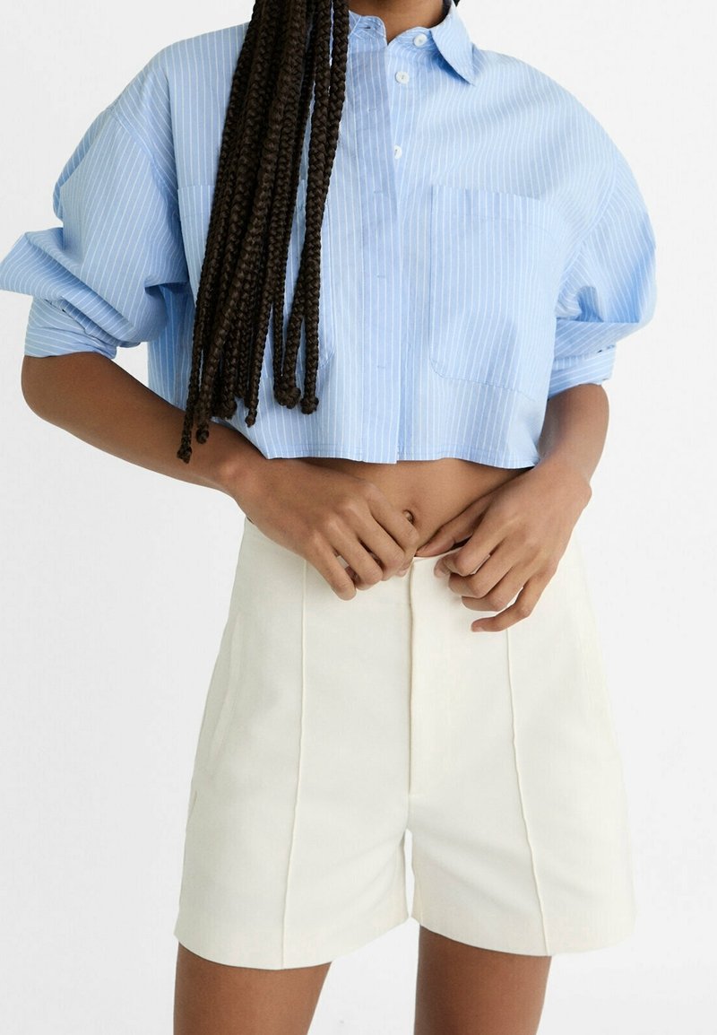 Camicia accorciata a righe blu chiaro con maniche corte e colletto abbottonato, abbinata a pantaloni corti bianchi a vita alta con pieghe.