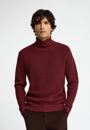 Uomo con capelli scuri e ricci che indossa un maglione a collo alto a coste color bordeaux e pantaloni scuri, in piedi con le mani in tasca davanti a uno sfondo neutro.