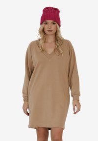 Beżowa sukienka z długim rękawem i dekoltem w kształcie V, w luźnym kroju. Zestawiona z teksturowaną fuksjową czapką roboczą.