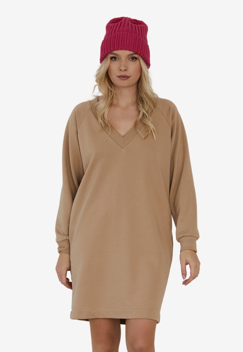 Beżowa sukienka z długim rękawem i dekoltem w kształcie V, w luźnym kroju. Zestawiona z teksturowaną fuksjową czapką roboczą.