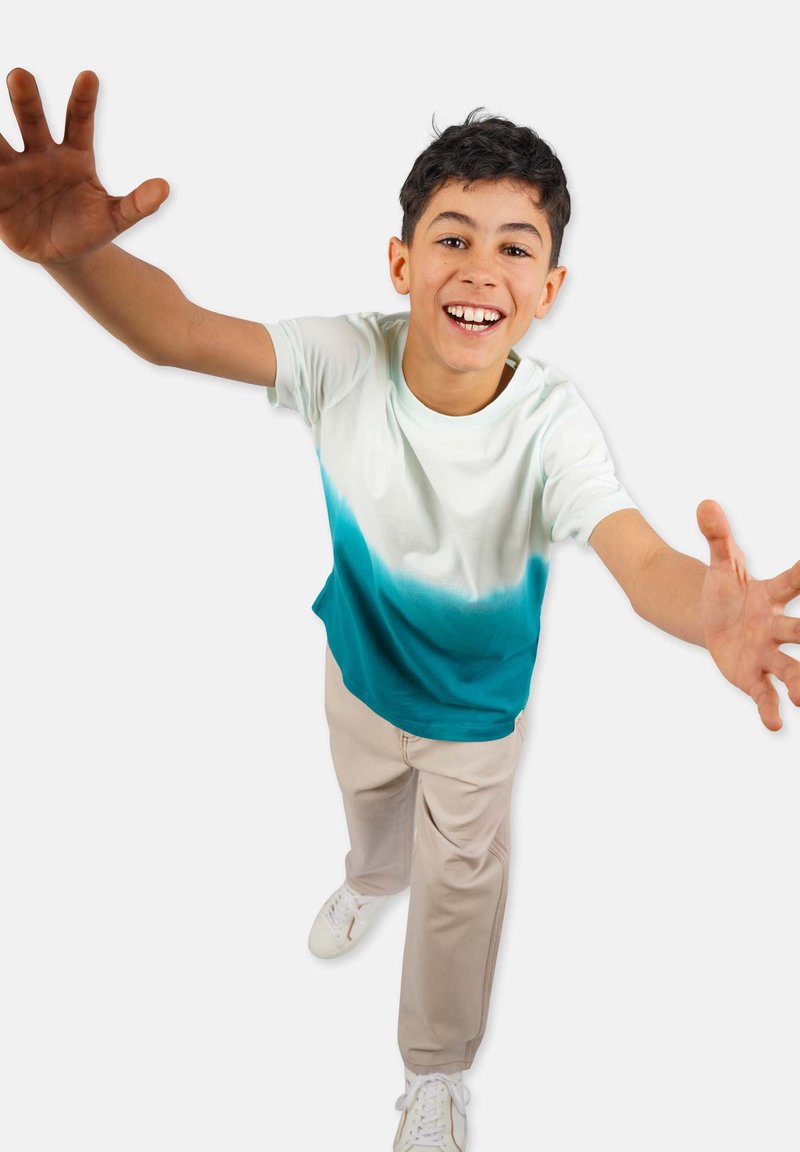 Short-sleeve t-shirt featuring a gradient from white to teal. Paired with beige trousers and white sneakers. Hands outstretched invitingly.