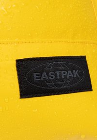 Gult vattentätt tyg med droppar, med en svart Eastpak-logotyp patch sydd på ytan. Texturerad finish, klar färg.