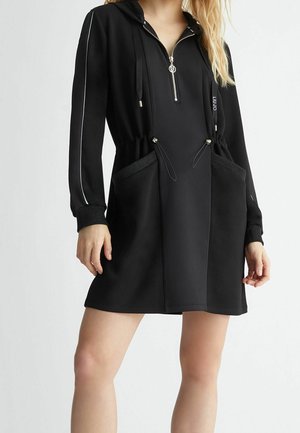 Femme portant une robe noire décontractée à manches longues avec fermeture éclair devant, capuche et taille à cordon avec poches.