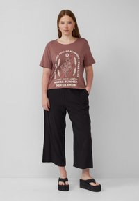Braunes Grafik-T-Shirt mit Rundhalsausschnitt und weißem Text, kombiniert mit weiten schwarzen Hosen und schwarzen Plateausandalen. Einfacher, lässiger Schnitt.