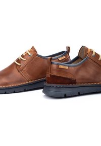 Zapatos de cuero marrón con acentos en color beige y suelas de goma gris. Las características incluyen cordones redondos, detalles de costura y etiquetas con el logo en los costados.