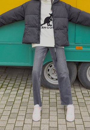 Person iført en sort dunjakke, beige sweatshirt med kengurulogo, grå jeans og hvide sneakers stående på flisebelagt fortov.
