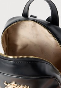 Mochila negra de cuero sintético con una cremallera dorada. El interior cuenta con un forro beige grabado con el nombre de la marca "Guess girl".