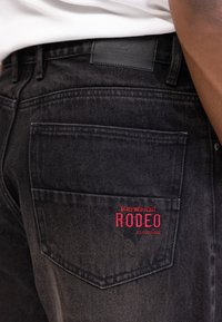 Crne traperice s izblijedjelom teksturom, sadrže stražnji džep s crvenim izvezenim logotipom "REDEFINED REBEL RODEO".