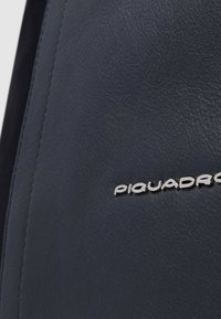 Bolso de cuero azul marino con textura suave, que presenta un logo plateado "PIQUADRO" en el lateral, mostrando un diseño minimalista y detalles de costura.
