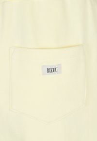 Lichtgele katoenen stof met een achterzak waarop een gestikt label staat met de tekst "BIZUU." De zak heeft een rechthoekige vorm en een gladde textuur.