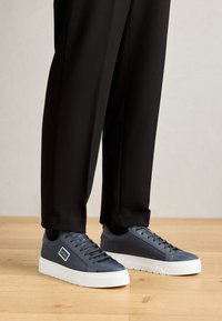Dark blue sneakers with a textured upper, white rubber sole, and minimalistic design, paired with black tapered pants on a wooden floor.