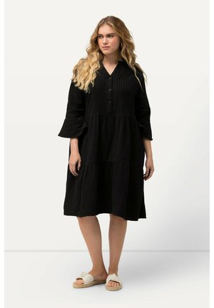 Robe en coton noir avec un devant à boutons, un tissu texturé, des manches évasées et un design à volants. Associée à des sandales de couleur claire.