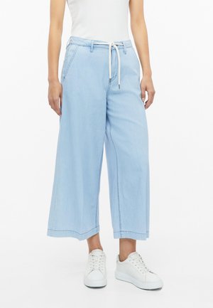 Persoon met wijde, lichtblauwe cropped jeans met een witte trekkoordriem, een wit mouwloos topje en witte sneakers, staat rechtop.