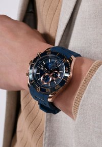 Ceas cu bratara din cauciuc albastru, cu carcasă rotundă de culoare auriu, cadran negru, accente aurii și trei sub-diale pentru cronograf. Include marcaje de oră îndrăznețe.