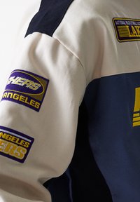 White and navy jacket with textured fabric, featuring embroidered yellow and purple logos: "Los Angeles Lakers" and "National Basketball Association."