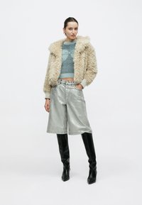 Beiger Kunstpelzmantel, blaue Strickjacke, metallisch silberne Shorts und schwarze kniehohe Stiefel. Das Model steht mit den Händen in den Taschen vor einem schlichten Hintergrund.