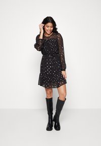 Robe noire transparente avec motif de pois dorés, taille cintrée et ourlet volants, associée à des bottes noires montantes. Manches longues avec poignets froncés.