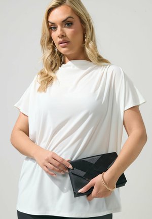 Lyshåret kvinde i hvid bluse med korte ærmer holder sort kuvertclutch, bærer gyldne hoop-øreringe og armbånd mod ensfarvet baggrund.