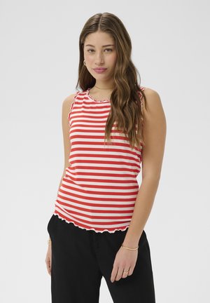 Jeune femme aux longs cheveux ondulés portant un haut sans manches rayé rouge et blanc et un pantalon noir, debout devant un fond gris uni.